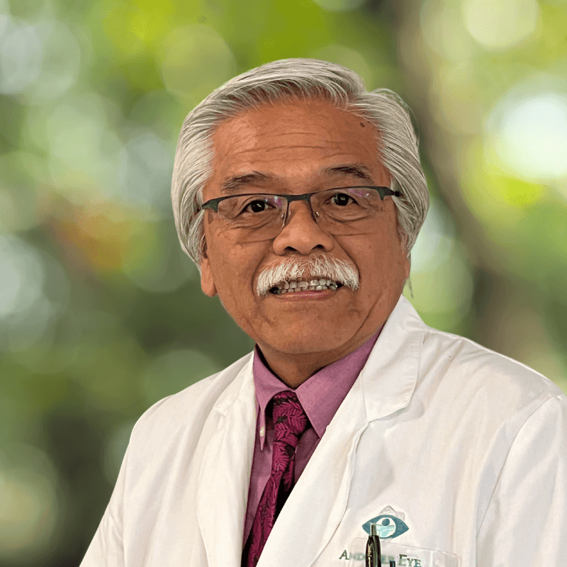 Man with gray hair and glasses in a white coat, smiling with a blurred green background.