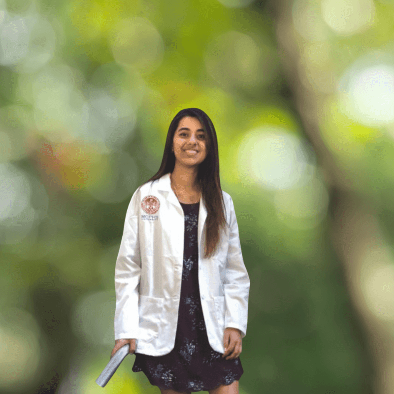 Person in a white medical coat holding a book, standing outdoors with a blurred green background.