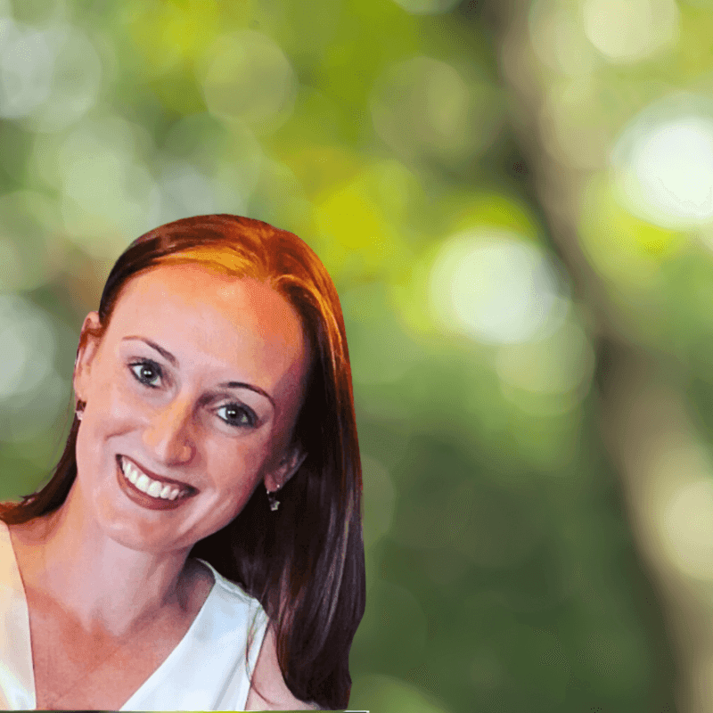 Smiling woman with brown hair standing outdoors with blurred green foliage in the background.