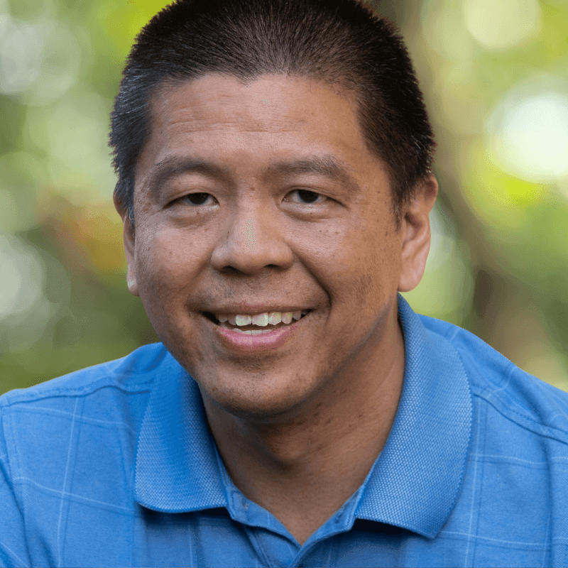 Close-up of a person wearing a blue collared shirt with a green and yellow blurred background.